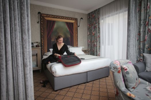 Femme déballant sa valise sur un lit dans une chambre d'hôtel élégante avec des rideaux fleuris.