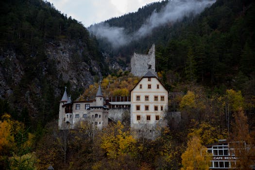 Château médiéval entouré de montagnes et de forêts, avec des couleurs automnales et un ciel nuageux.