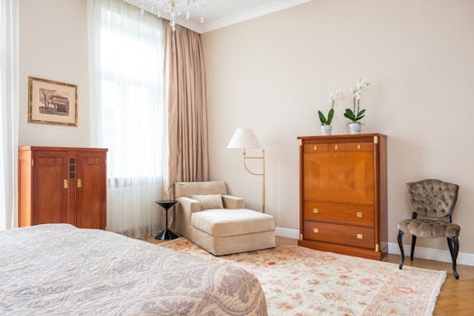 Chambre élégante avec lit, fauteuil, commodes en bois et plantes, dans un décor lumineux et raffiné.