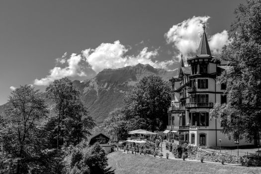 Château élégant entouré d'arbres, avec montagnes en arrière-plan et terrasses animées.